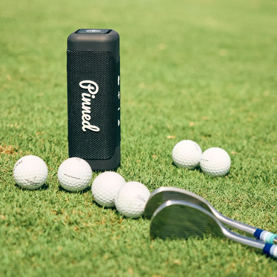 navy pinned golf speaker in grass with golf balls and golf clubs